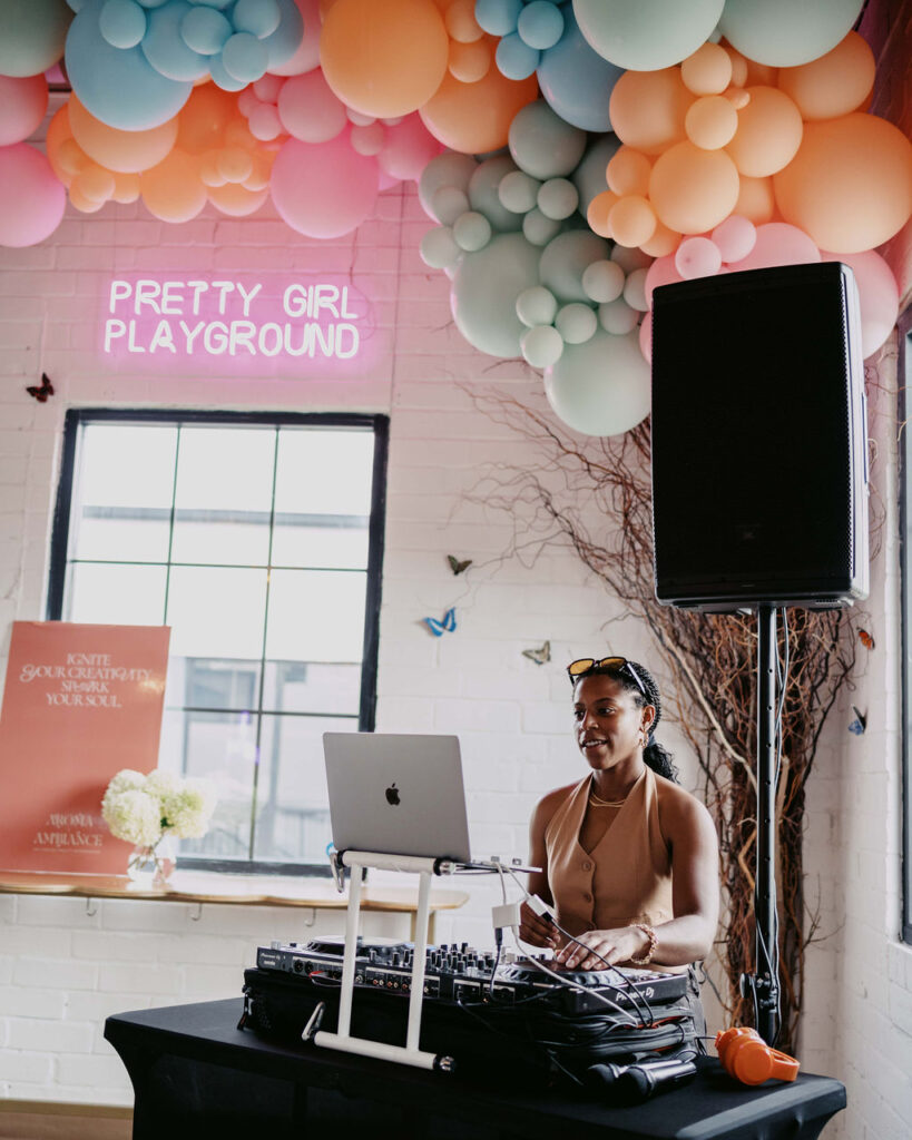 DJ performing at the Aroma & Ambiance event by Indigo Pruitt Designs, with colorful balloon decorations and a 'Pretty Girl Playground' neon sign in the background.