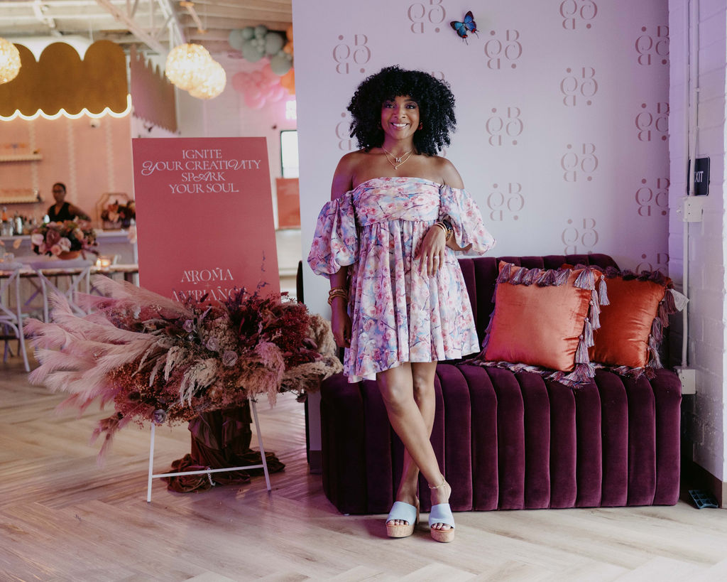 Marie Cloud of Indigo Pruitt Designs standing in front of a styled backdrop at the Aroma & Ambiance event, featuring elegant floral arrangements and a welcoming sign that reads 'Ignite Your Creativity, Spark Your Soul