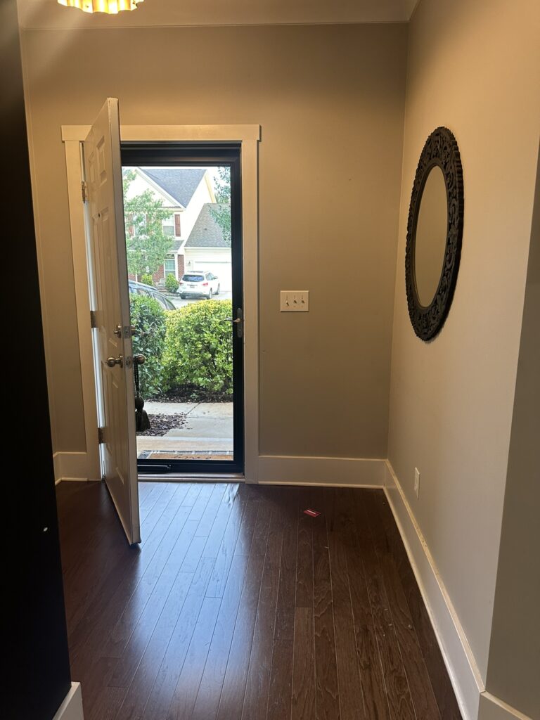 Entryway with an open front door leading to a view of the neighborhood. The space features hardwood flooring, neutral walls, a round decorative mirror on the right wall, and a ceiling light fixture. Sunlight illuminates the area, highlighting a welcoming ambiance.