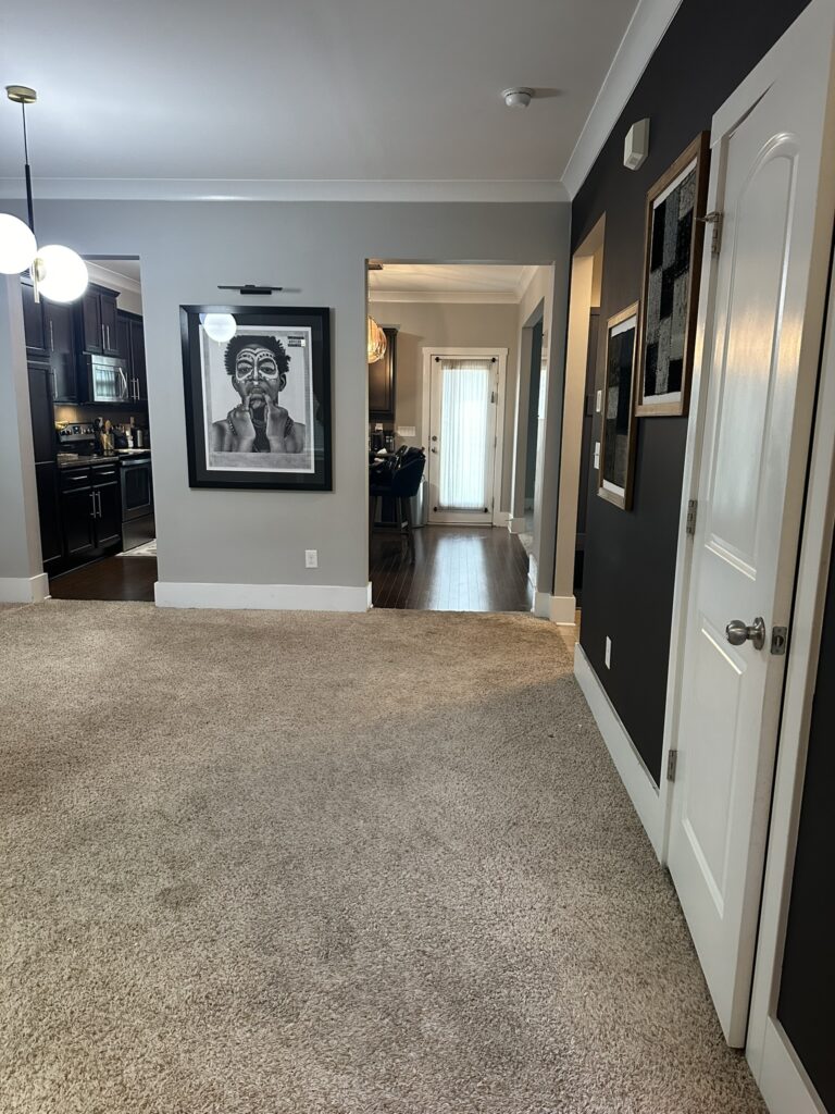 Dining room with a neutral carpeted floor and dark accent walls. The room features framed artwork, including a prominent black-and-white portrait. The view extends to an adjacent hallway with hardwood flooring, leading into a kitchen area with dark cabinetry and a glass door at the back.